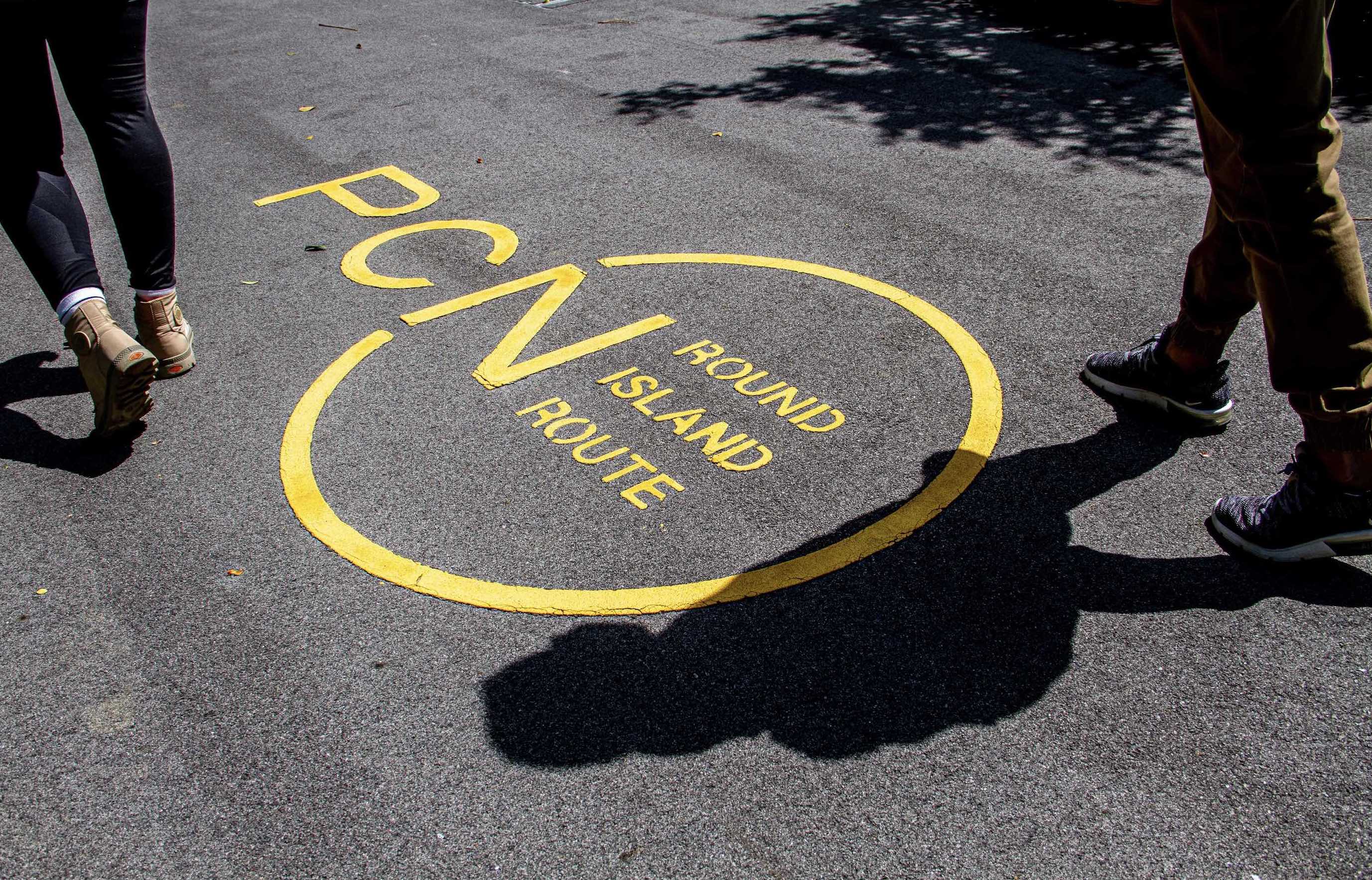Two people walking on a path with the text "PCN ROUND ISLAND ROUTE" inside a yellow circle on the ground.